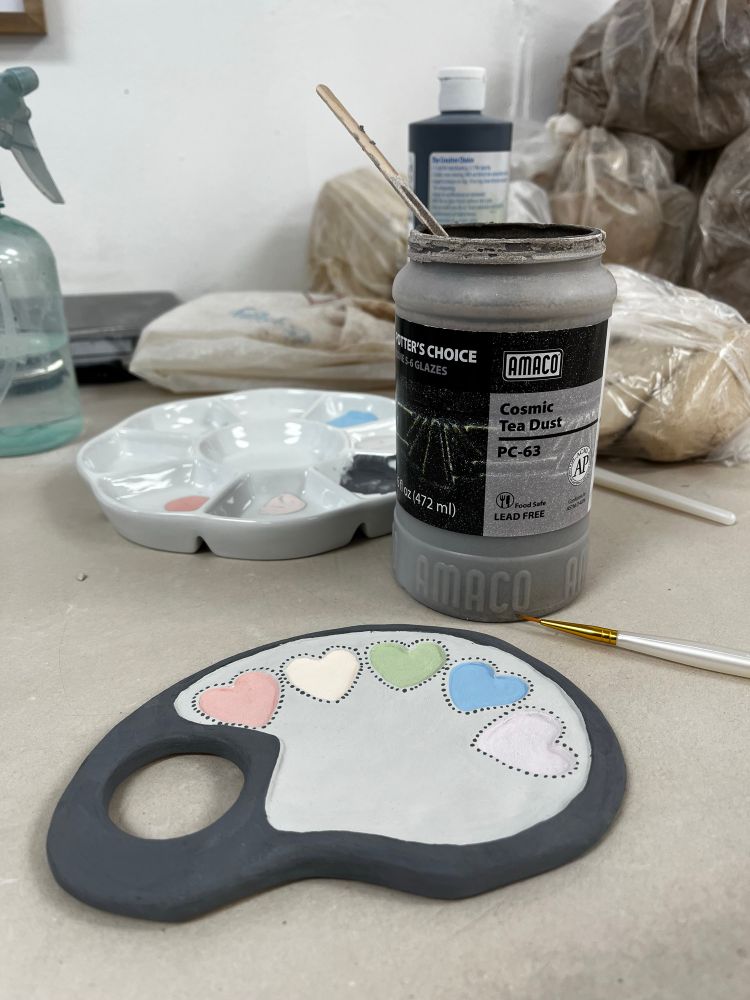 A paint palette in progress with five heart-shaped paint wells surrounded by grey and black. In the background is a paint palette and an open bottle of Amaco’s Cosmic Tea Dust glaze, and various bags of clay.