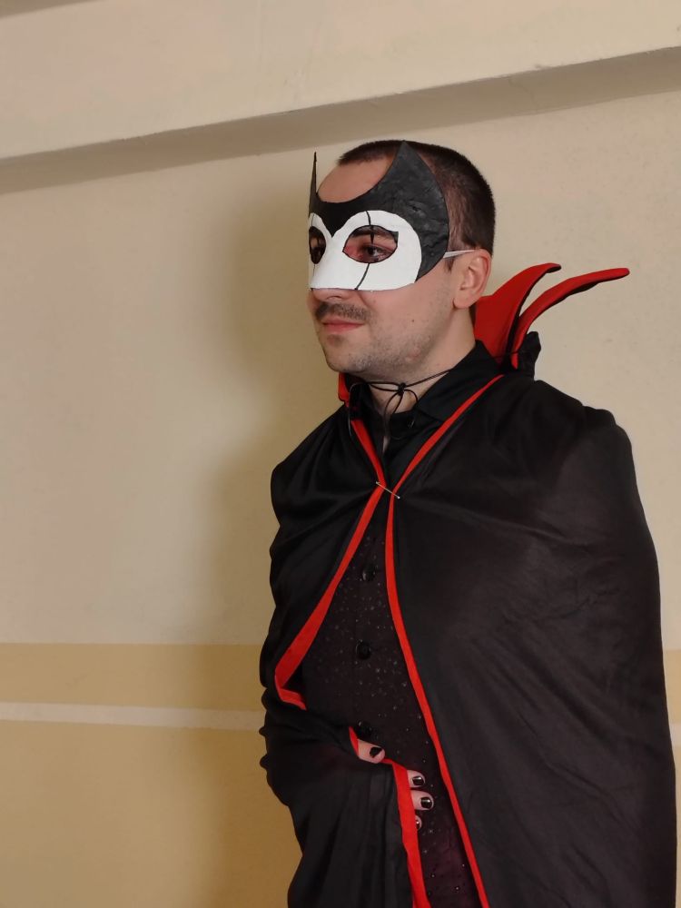 Photo of the same person more up close. They are wearing a black mask with white accentuation covering the eyes and a black cape with red lining + black clothes underneath.
