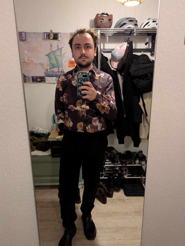 Spiegelselfie von einer weißen Person mit verwuschelten blond gefärbten und dunkelbraunen Haaren. Die Person trägt ein dunkles Hemd mit einem Blumenmuster und eine schwarze Hose.