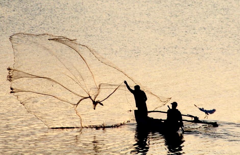 Silhouette of 2 fisherman on a small simple wooden boat floating on yellowy sunset-tinged water One is standing casting a large net the other is sitting down rowing. The bird has its wings spread about to land back on the deck. 