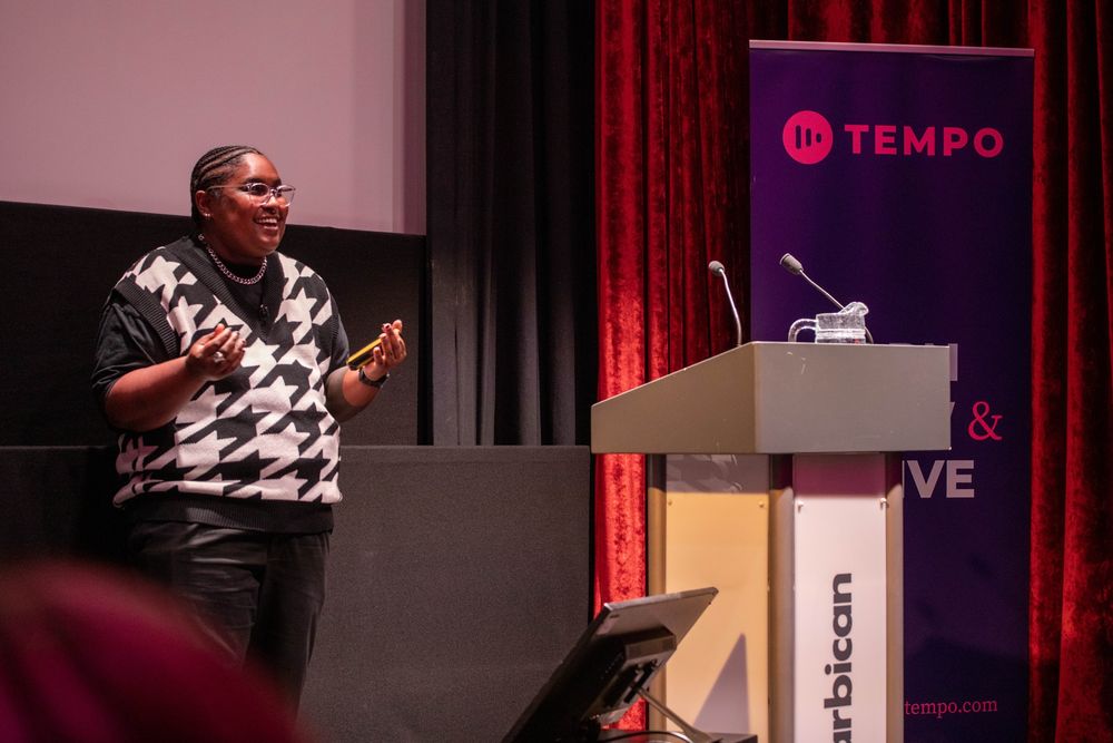 A stage and lecturn with someone in a black and white top speaking and gesturing 