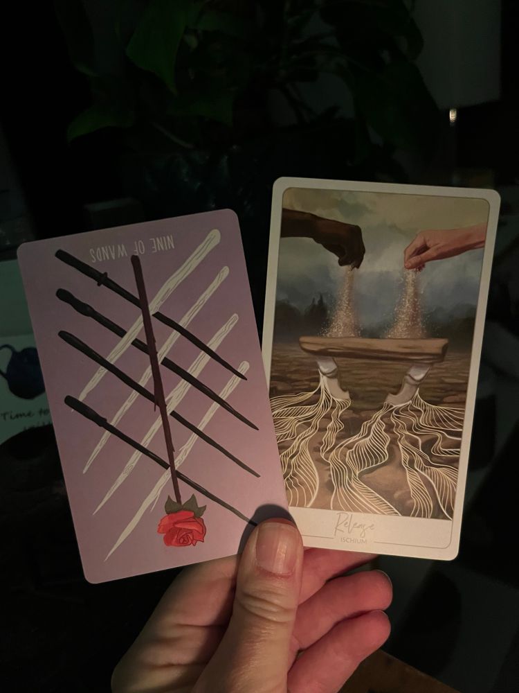 A white hand, illuminated by candle in the early morning light, holding two cards. The one on the left the Tarot card 9 of Wands reversed, the one on the right an oracle card: Release (ischium).