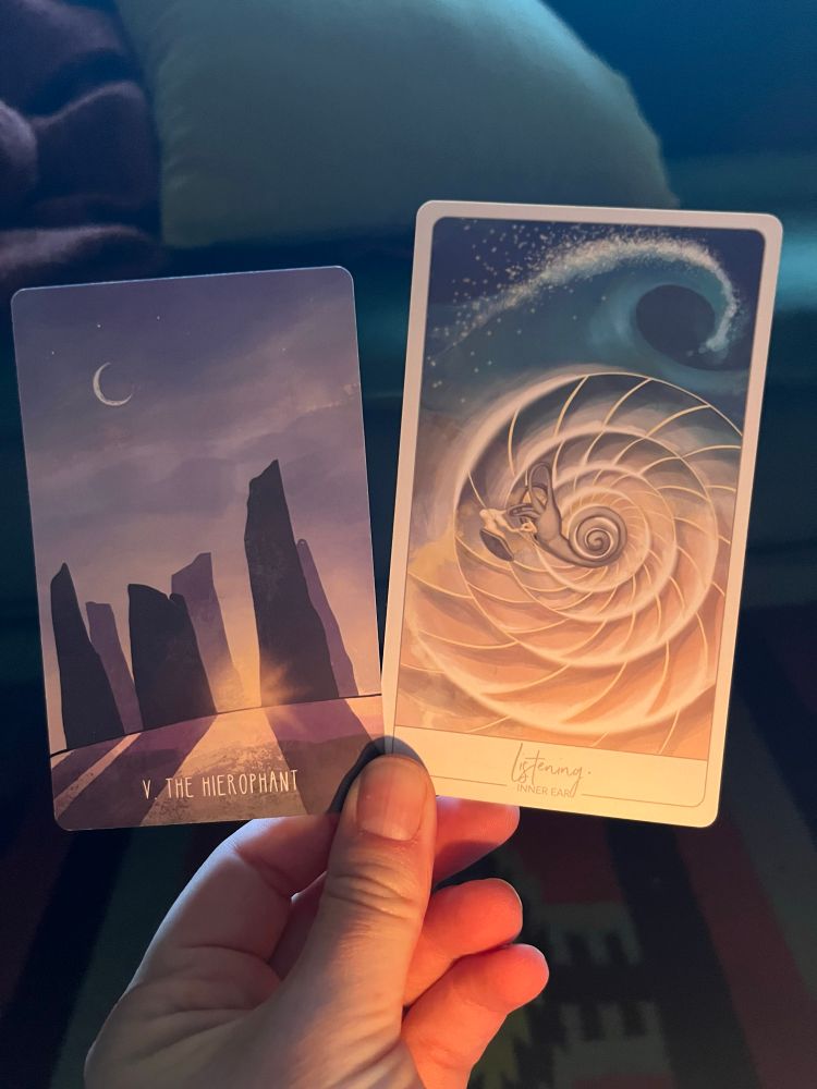 A white hand, illuminated by candle in the early morning light, holding two cards. The one on the left the Tarot card The Hierophant, the one on the right an oracle card: Listening (Inner Ear).