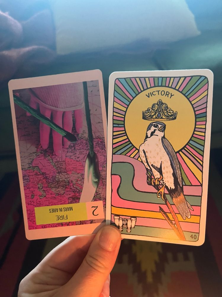 A white hand, illuminated by candle in the early morning light, holding two cards. The one on the left the Tarot card 2 of Wands reversed, the one on the right an oracle card: Victory.