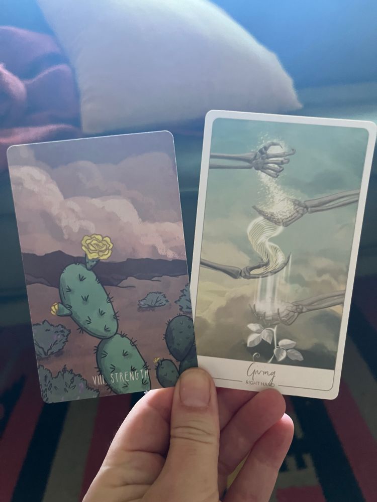 A white hand, illuminated by candle in the early morning light, holding two cards. The one on the left the Tarot card Strength, the one on the right an oracle card: Giving (right hand).