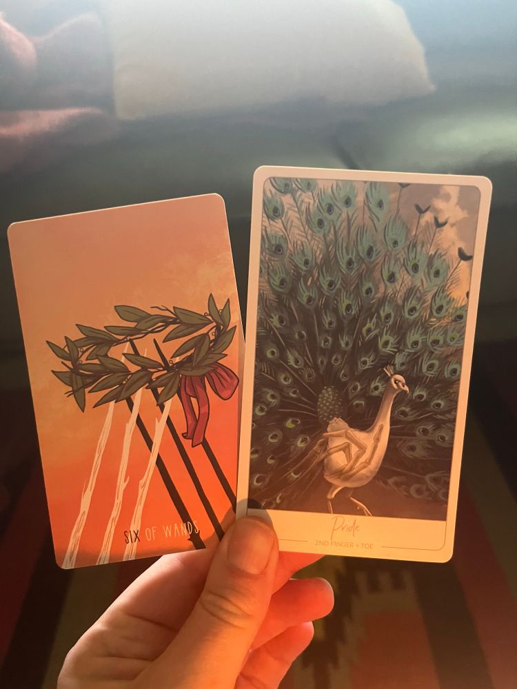 A white hand, illuminated by candle in the early morning light, holding two cards. The one on the left the Tarot card 6 of Wands, the one on the right an oracle card: Pride (2nd finger and toe).