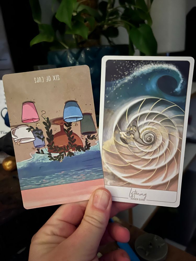 A white hand, illuminated by candle in the early morning light, holding two cards. The one on the left the Tarot card 6 of Cups reversed, the one on the right an oracle card: Listening (inner ear).