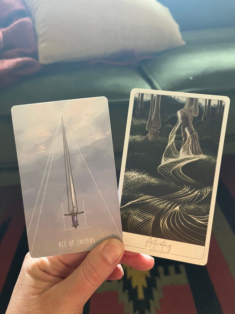 A white hand, illuminated by candle in the early morning light, holding two cards. The one on the left the Tarot card Ace of Swords, the one on the right an oracle card: Activating (right foot).