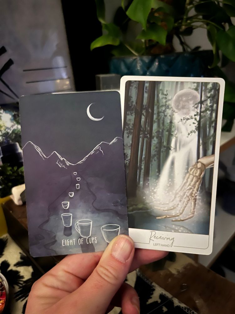 A white hand, illuminated by candle in the early morning light, holding two cards. The one on the left the Tarot card 8 of Cups, the one on the right an oracle card: Receiving (left hand).