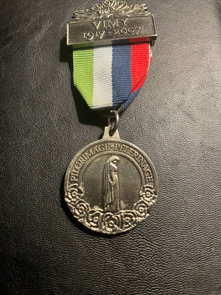 Pilgrimage medal from the 90th anniversary of the Battle of Vimy Ridge. Silver, metal bar at the top with the dates 1917 - 2007. The ribbon has four stripes: from left to right, green, white, blue, and red. They represent the four divisions of the Canadian army fighting together for the first time. On the medal itself is an image of Young Canada grieving for her soldiers. On the edge, at the top it says “Pilgrimage” in English and French. At the bottom there are poppies. 