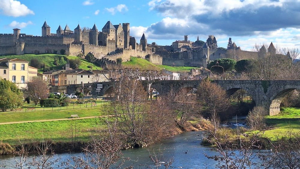 A fairytale-like stone castle inside a walled city sits atop a hill of varying greens under a light blue sky with a few large puffy clouds. In the lower half of the image, an old bridge crosses a small river that runs right towards the viewer.