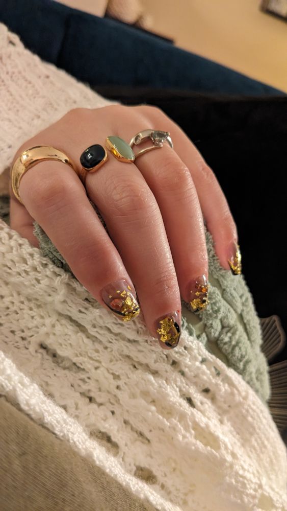 Photo of a hand with gel nails. The design is abstract, gold, black, and copper gems