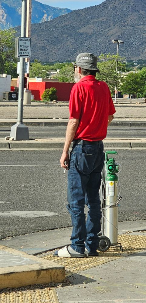 Man in red shirt and jeans waiting to cross  the street toting a oxygen tank in one hand and a lit cigarette in the other.