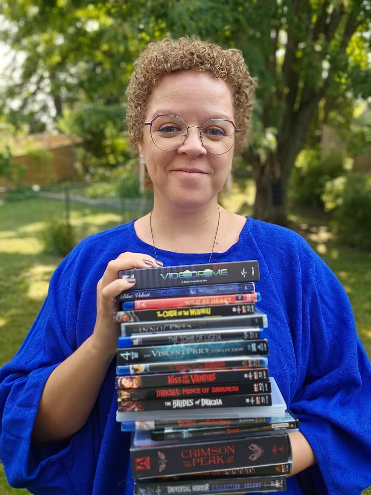 A picture of me smiling while holding a big stack of horror movies as research for my upcoming book.