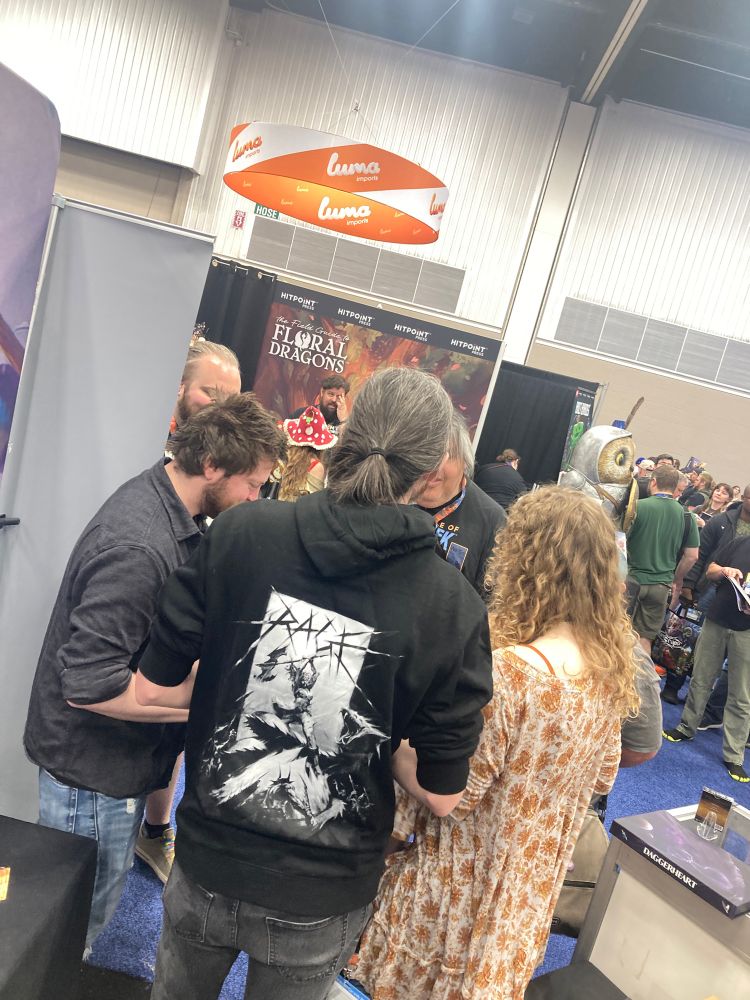 photo of the backs of spenser starke, shaun ellis, and rowan hall while they sign stuff in a crowded convention hall.