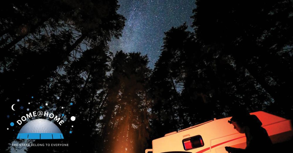 An individual sitting outdoors among trees in front of a fire pit next to a camper van, under a night sky filled with stars.