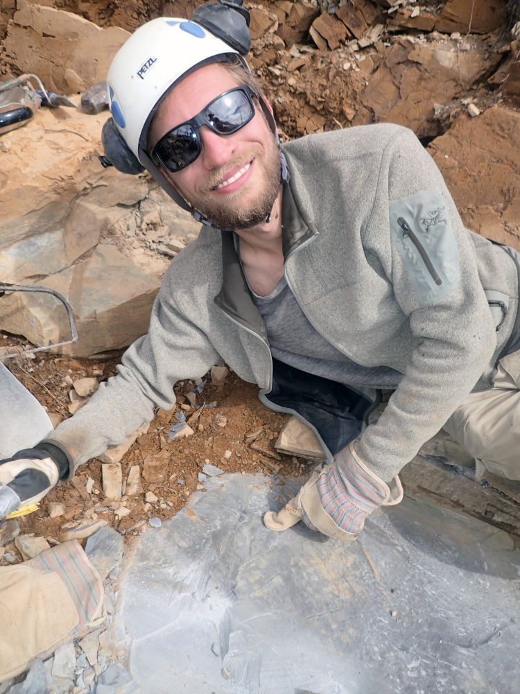 Manitoba Museum Curator of Palaeontology and Geology, Dr. Joe Moysiuk, wearing sunglasses, a hard hat, and ear protectors, posing next to an uncovered fossil in a piece of rock.