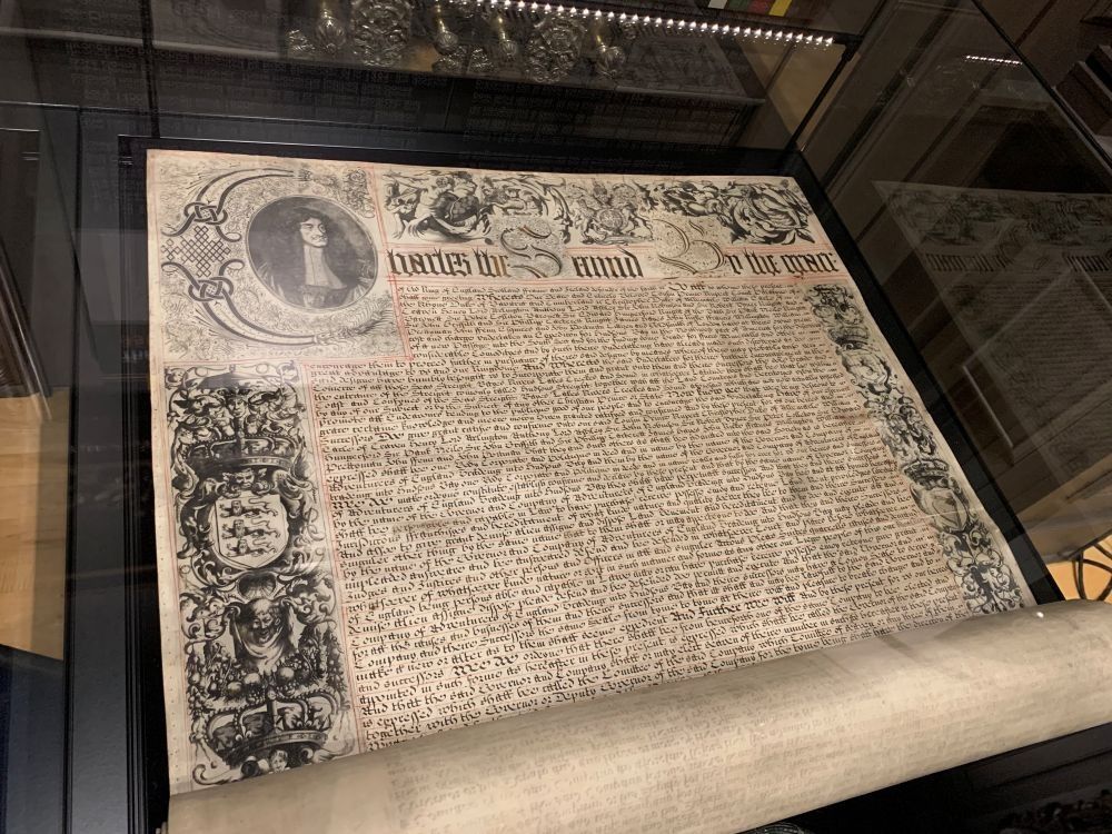 The HBC Charter on display in a glass case. A wide piece of parchment, still rolled at the bottom with intricate designs in the margins and the wording of the charter written with flourishes through the body of the page.