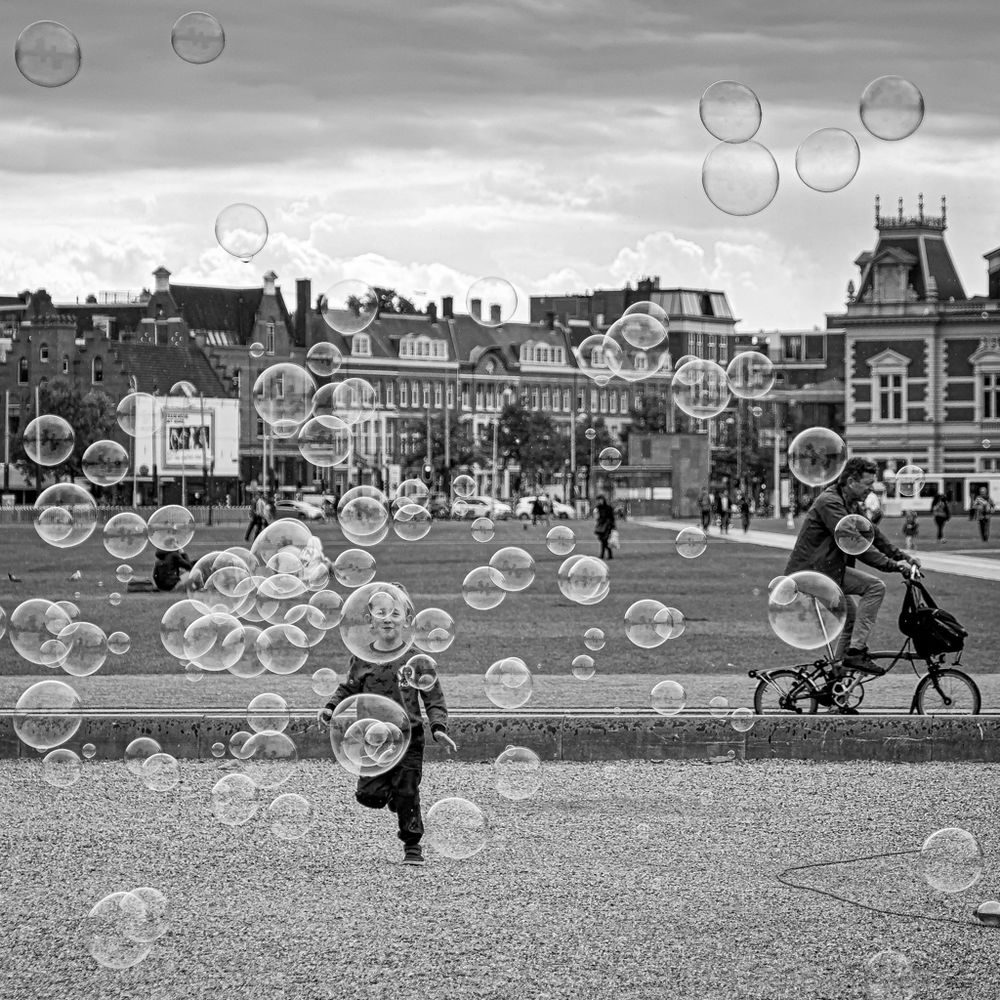 B&W image of a young boy runs through bubbles in Amsterdam, NL, one of which frames his face. 