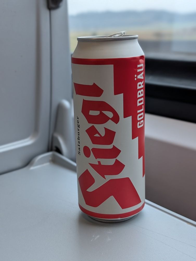 A 0.5l red and white somewhat retro looking Stiegl beer can on the fold-out seat-back table. One can see the rolling hills of Bavarian Swabia out the train window in the background.