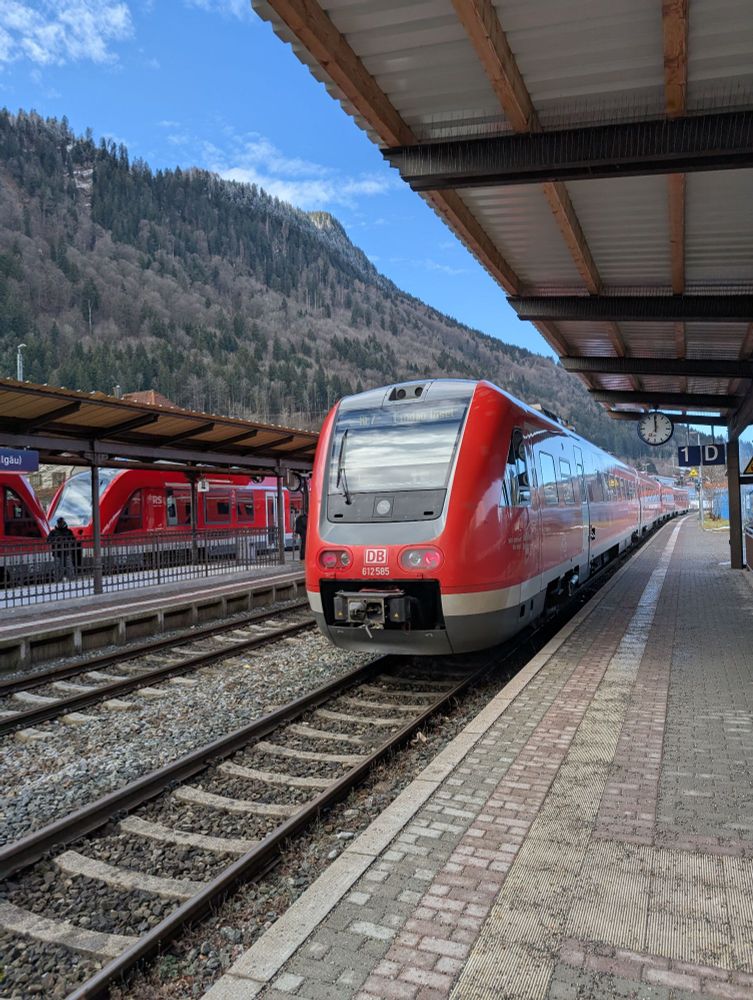 A DB Class 612 diesel multiple unit pulling out of Immenstadt train station. The train is painted in DB's standard bright red. On the left and in the background are the hills of the pre-Alp area. On the right, the covered platform with a nice analog clock hanging from the roof.
