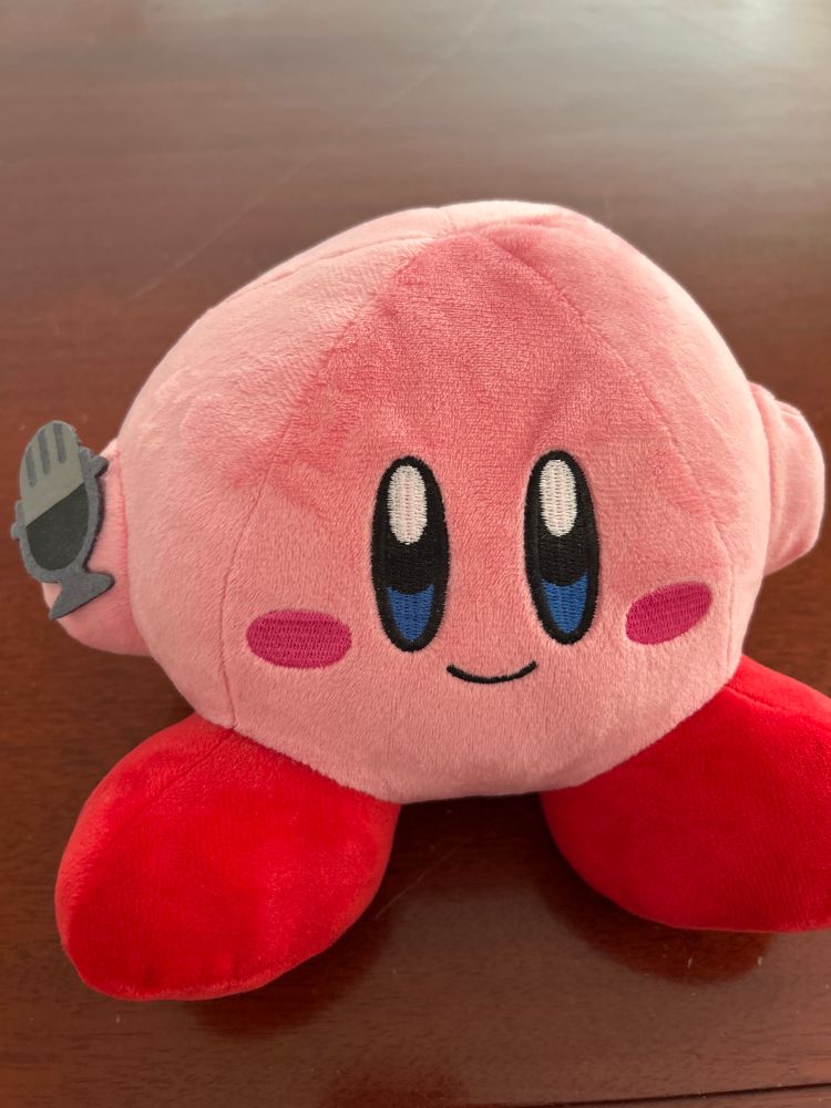 A cute little plush Kirby Bluetooth speaker holding a silver microphone.