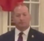 An image of a balding white man with a dark suit jacket, white shirt, and red tie -- presumably a member of the Secret Service -- with a look of disappointment on his face as he watches the richest man in the world play with silverware like a child.
