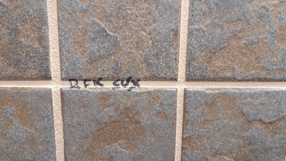 A tile wall with a hard to read "RFK SUX" written in Sharpie on the grout. 