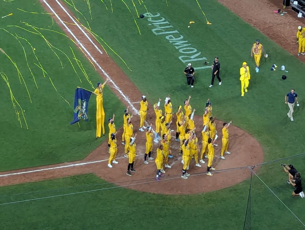 The Savannah Bananas players in yellow uniforms dancing over home plate with yellow streamers falling behind them
