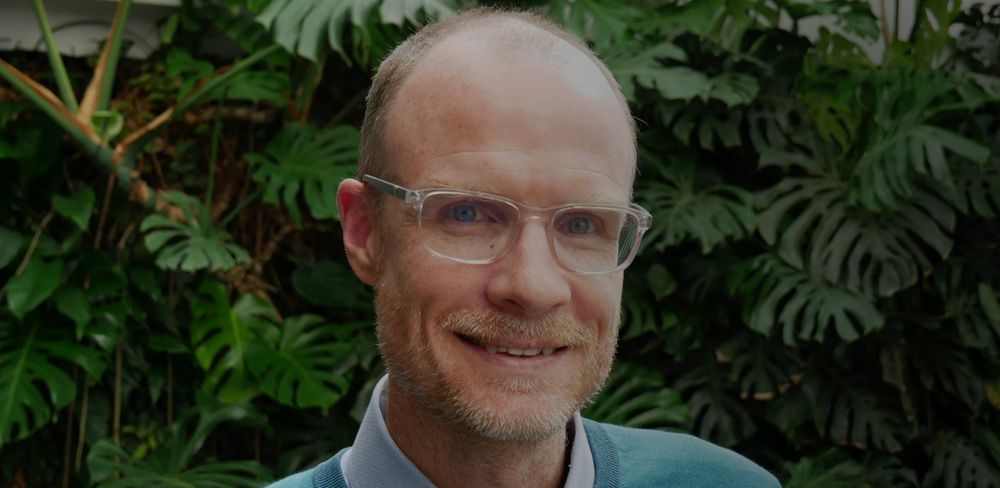 Professor Chris Sandbrook photographed in front of a lush backdrop of large, dark green tropical foliage.