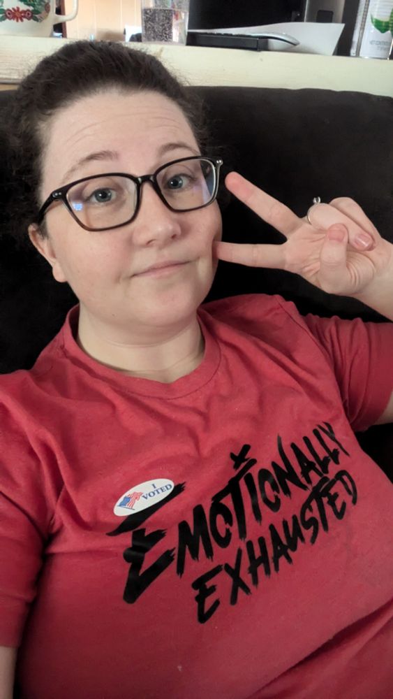 Skw3rlch4n is posing with a peace sign next to her cheek. She is wearing a red shirt that says Emotionally Exhausted, with a 'I Voted' sticker on.