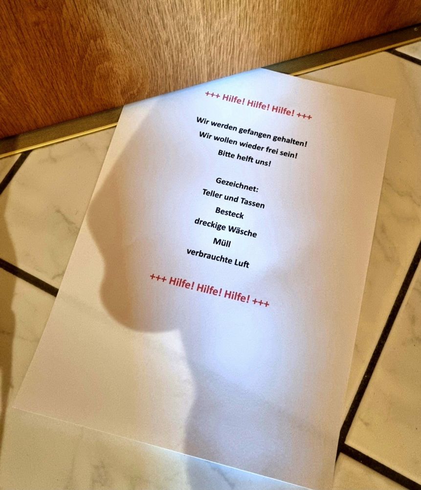 Blatt Papier, welches unter der Zimmertür hervorschaut, mit der Aufschrift: Hilfe, wir werden gefangen gehalten! Wir wollen wieder frei sein! Bitte helft uns! Gezeichnet: Teller und Tassen, Besteck, dreckige Wäsche, Müll, verbrauchte Luft