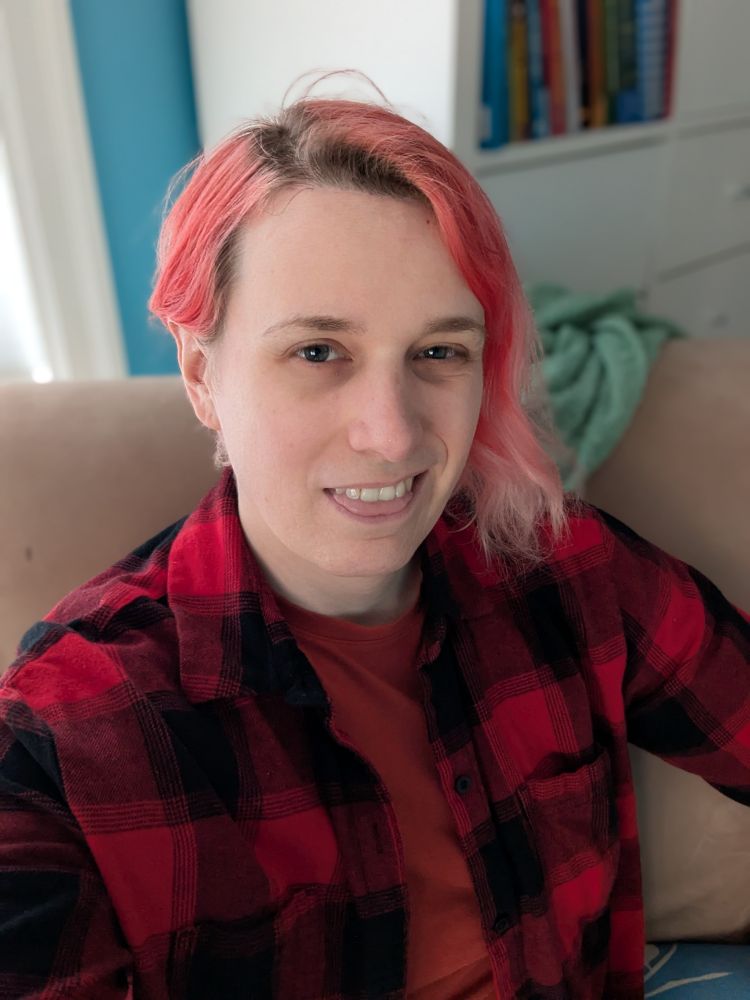 Selfie. I am making a smirking smiling face with my tongue between my bottom lip and my top row of teeth. I am looking towards the camera and wearing a red shirt with a red flannel over top.