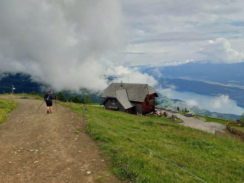 Eine Wanderin auf dem Weg vorbei an einer Wanderhütte - den Blick ins Tal gerichtet - auf einen See. Vor ihr zieht eine dichte Wolke entlang.