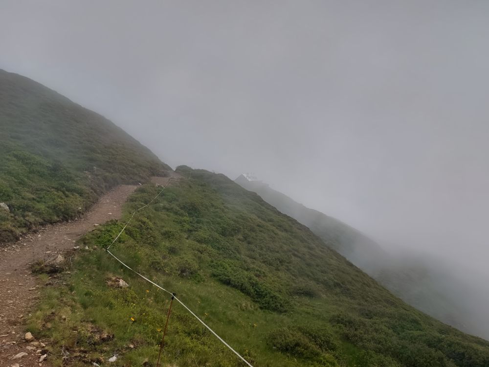 Ein Bergpfad im dichten Nebel. Durch den Nebel sind schwach die Konturen einer Berghütte erkennbar.