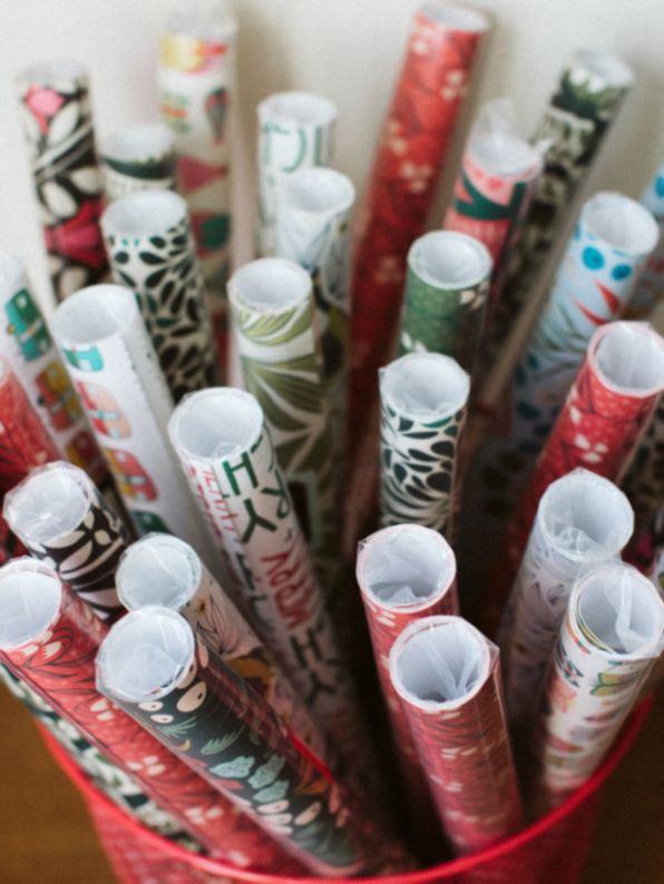 basket filled with festive rolls of wrapping paper designed by Rebecca Jane Woolbright, featuring colorful holiday patterns like snowflakes, trees, and florals — perfect for seasonal gifting and Christmas wrapping.
