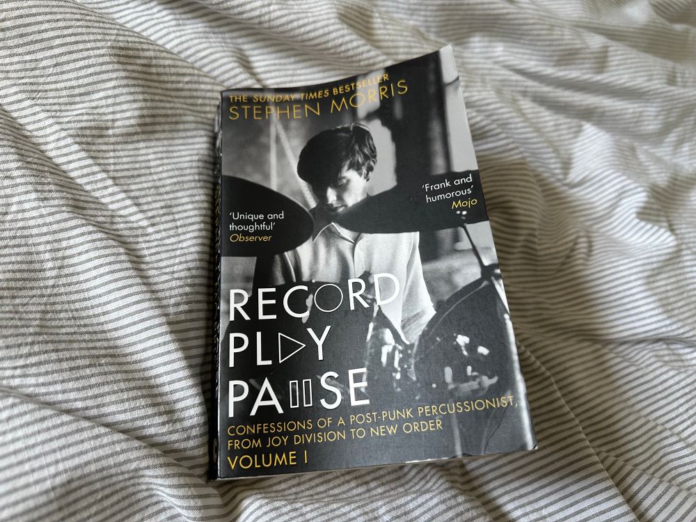 A paperback copy of “Record Play Pause” by Stephen Morris. The cover features a black & white photograph of the author seated behind a drum kit. The author’s name and book title are written in white and dark yellow fonts. 
