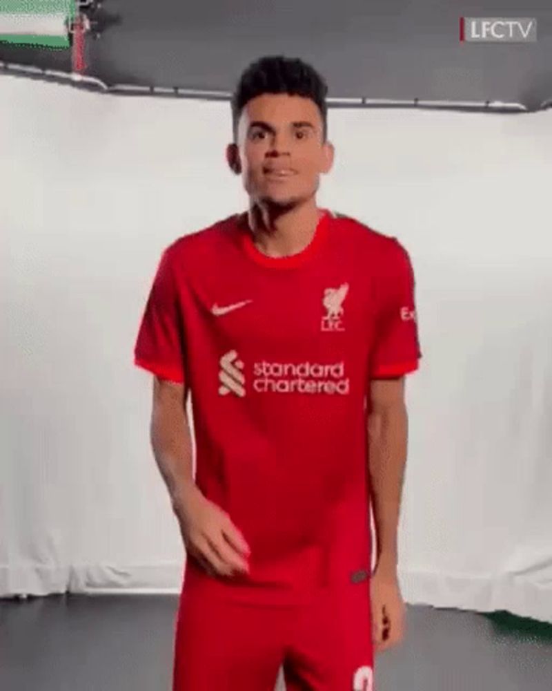 a man in a red soccer uniform is standing in front of a white wall and smiling .