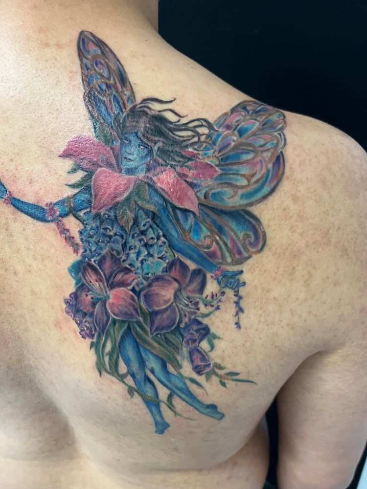 Tattoo of a blue female fairy in a dress of bell flowers and lily’s with butterfly wings.