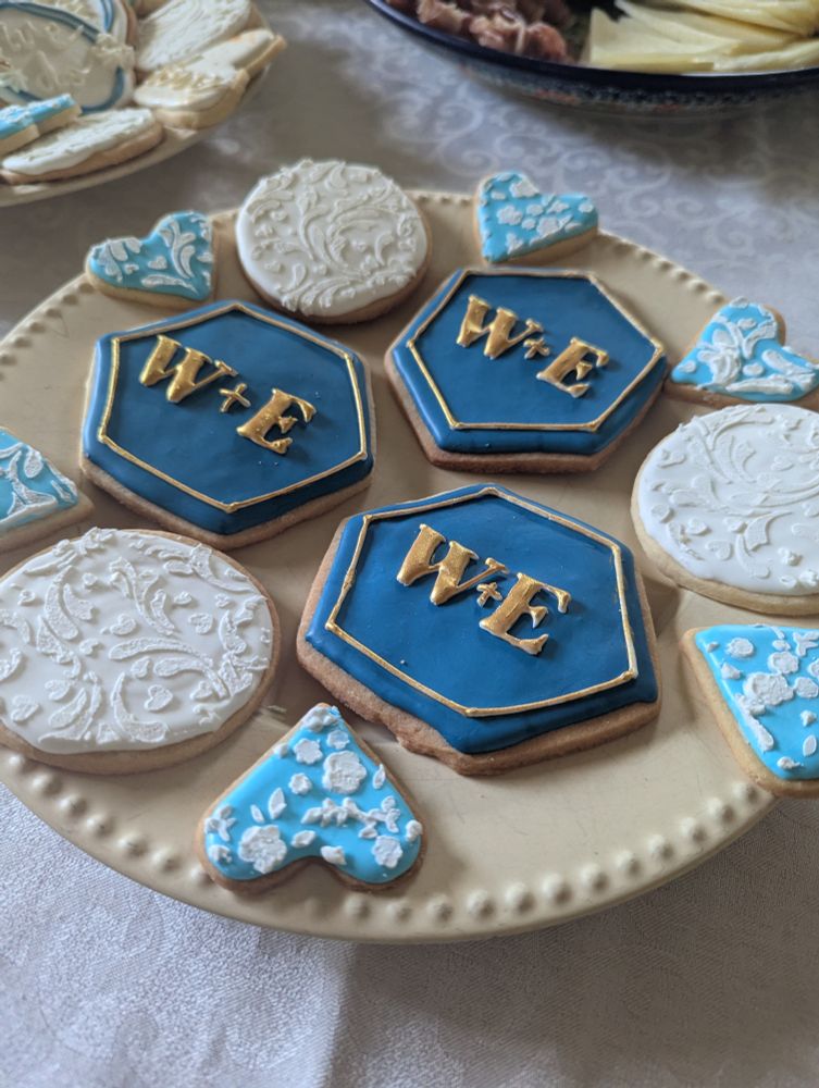 Blue, white and gold cookies on a plate. The main cookies are dark blue hexagons with W+E written in gold. These are surrounded by white circles stenciled with a floral pattern, and mini blue hearts stenciled in white. 