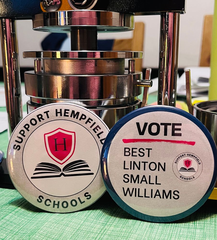 A button maker sits behind two campaign buttons. The left button says, “Support Hempfield Schools,” and displays a logo of an H inside a red shield over an open book. The right button has a blue background and displays the word, “VOTE” above the name of four Hempfield School District school board candidates - Best, Linton, Small, Williams.