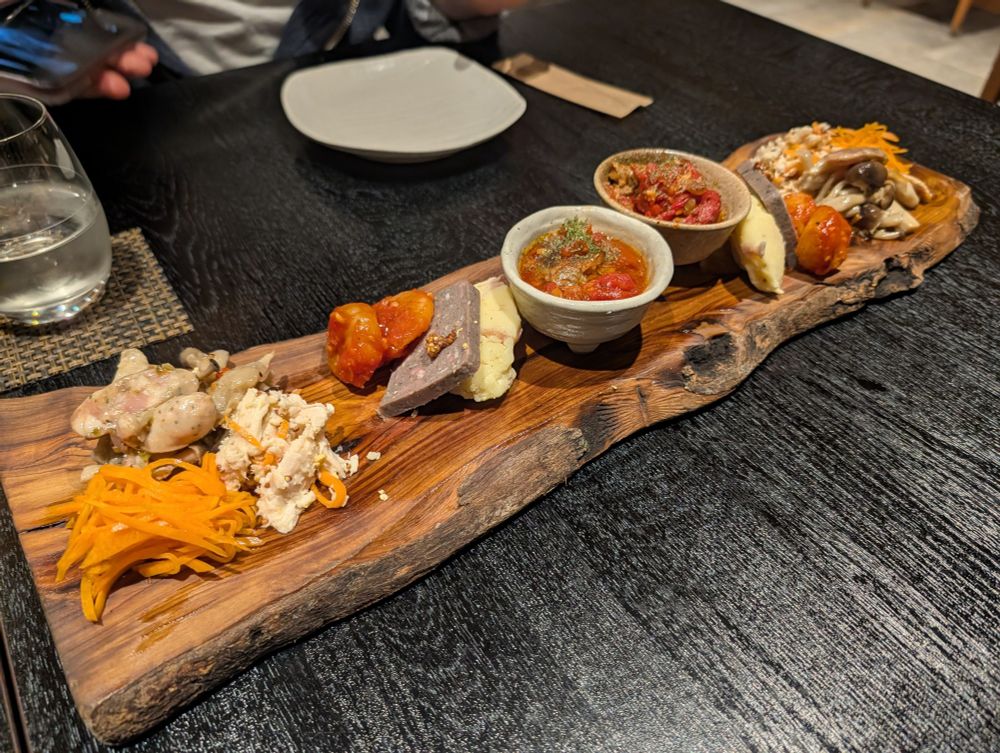 Photo of an appetizer tray displayed on a wooden slab. There's carrots, mushrooms, shrimp, chicken, potatoes, and a small cup of wagyu beef in a tomato sauce