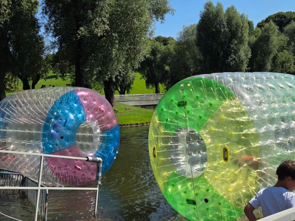 Das Bild zeigt zwei große, transparente Wasserkugeln, die teilweise in einem See liegen. Die Kugeln sind in verschiedenen Farben (rosa/blau und grün/gelb) gehalten. Im Hintergrund ist ein grüner Park mit Bäumen und einem kleinen Steg zu sehen. 