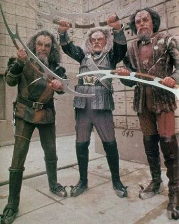 Promotional still from the television show Star Trek: Deep Space 9, Season 2 Episode 19 "Blood Oath" which guest stars three Klingon Characters (Kor, Koloth, and Kang) that had been featured in previous Star Trek iterations.

Photo Description: Three humanoid alien beings with large statures, big bumpy foreheads, and long salt-and-pepper to white hair stand in a row in a hieroglyph-laden sand stone courtyard holding up Klingon combat weapons called "bat'leths" in a triumphant manner. Their clothing mixes Viking, Mongolian, and Russian aesthetics with big belts and padded coats, and they all wear big black boots with curved horns jutting off the toe-end.