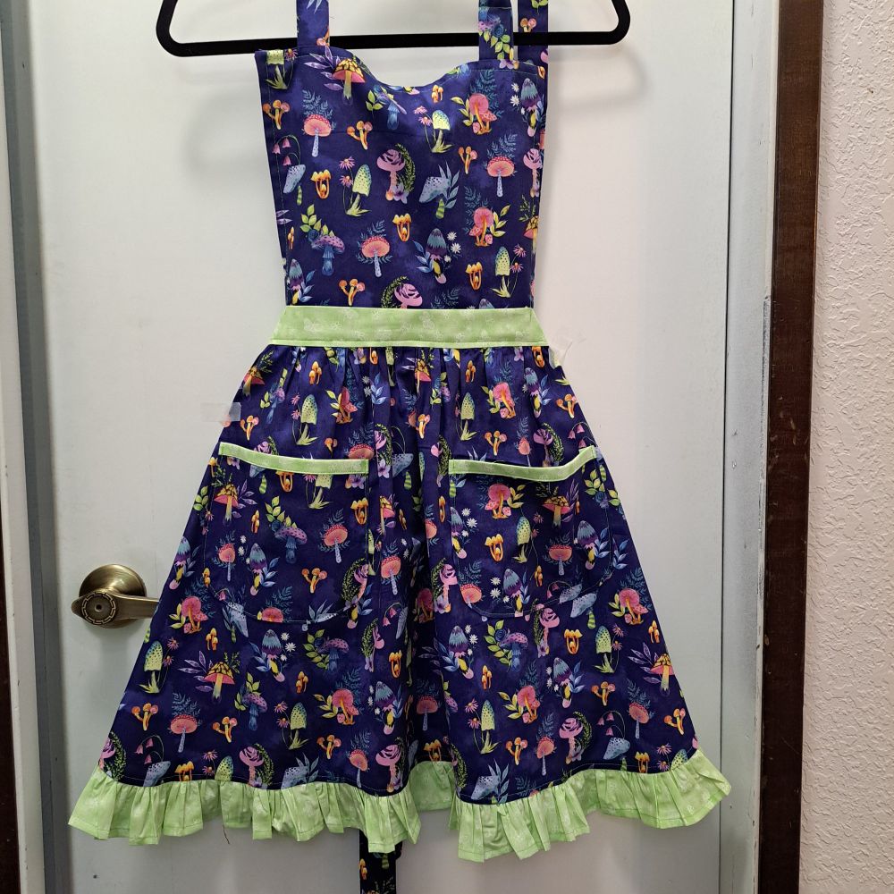 A dark blue apron with different neon pink, blue, orange, and green colored mushrooms. It has pockets highlighted with bright green trim. The same bright green is used on the waistband and ruffles at the bottom. It has adjustable neck and back ties. The cotton is machine washable.