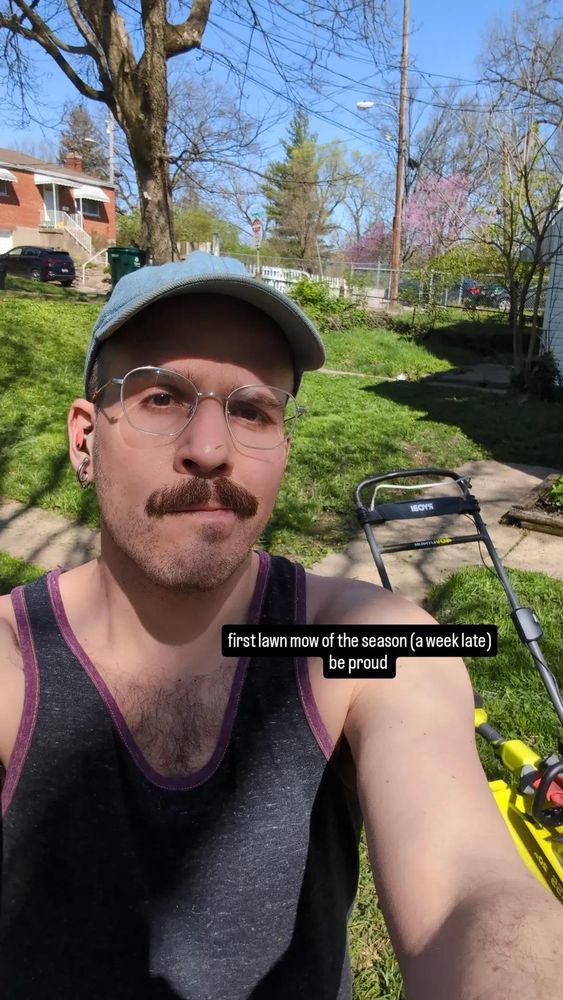 a thirsty man with a mustache in a blue ball cap and glasses stands in from of lawn equipment in a freshly mowed yard. pink flowers and new leaves can be seen in the background.