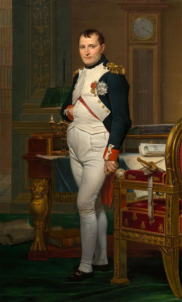 Portrait of Napoleon Bonaparte, standing in front of an ornate, gilded, desk and chair.
