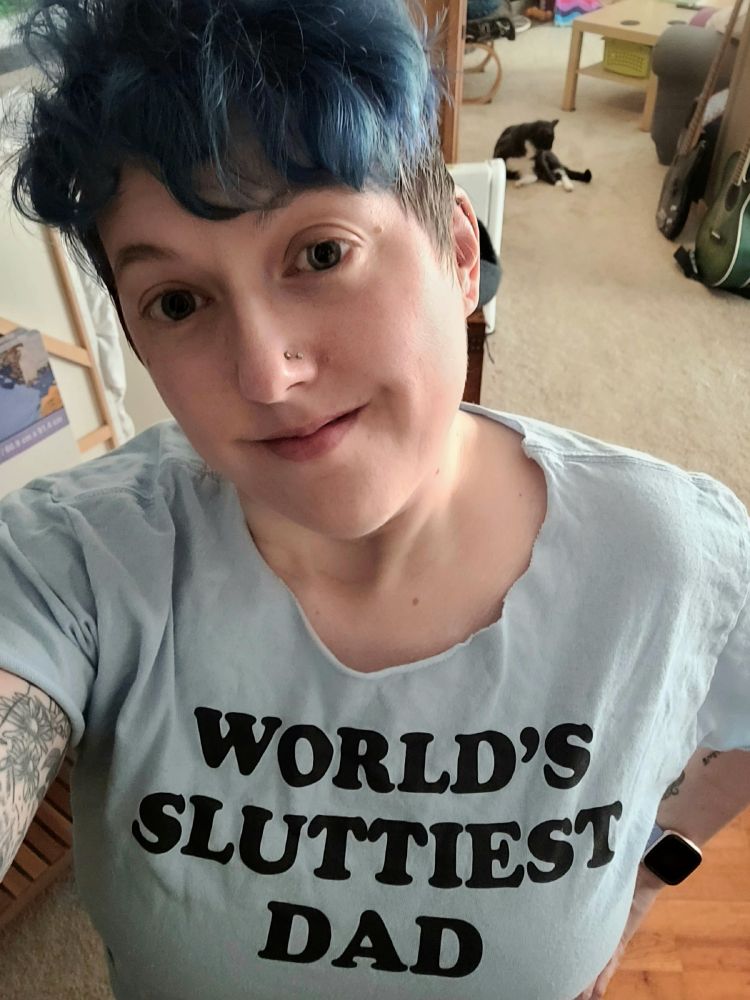 Selfie of a tired person with blue hair wearing a t-shirt reading "World's Sluttiest Dad"