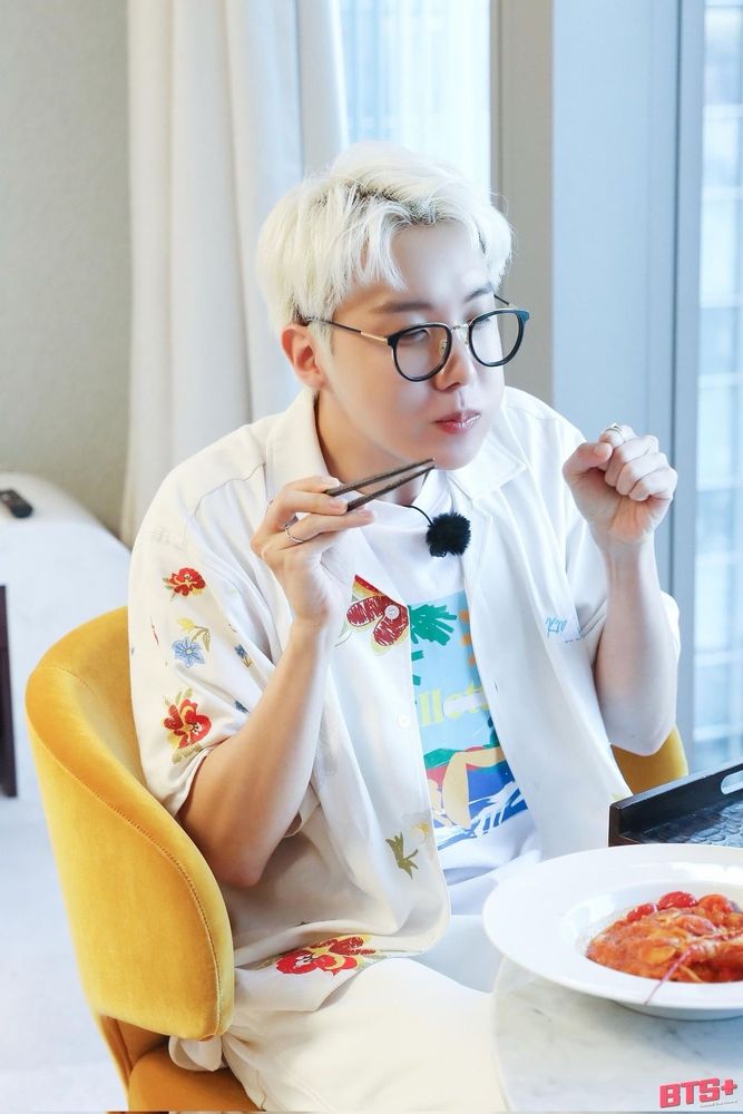 Hobi with blond/ white hair sitting on a yellow chair holding a pair of chopsticks making a little fist cause he’s so happy about his 