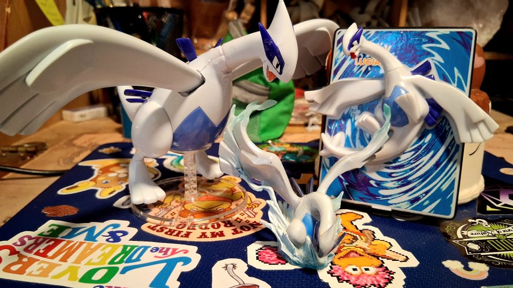 The photograph is of three figurines of the Pokémon Lugia in three different sizes, arranged on top of a laptop covered with many stickers.